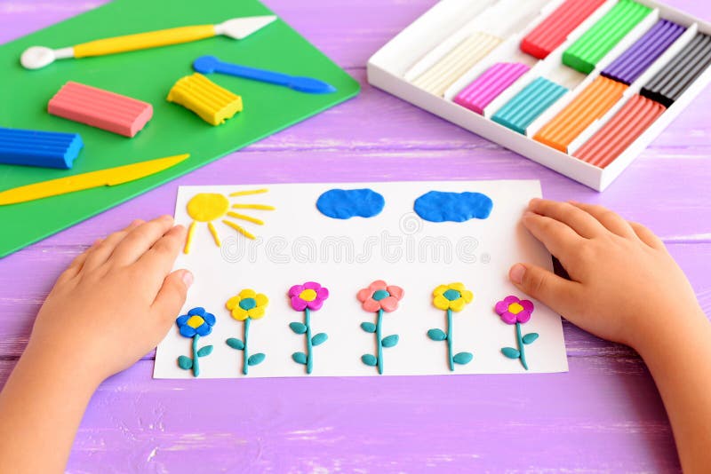 Child shows a card with plasticine flowers, sun and clouds. Supplies for children art crafts on wooden table. Modeling clay craft. Modeling clay crafts idea for stock photos