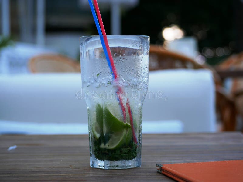 A glass of cold Mojito with ice is on the table on the summer veranda of the cafe. The glass with the tube is covered with droplet. S of condensate. Drink on a stock photo