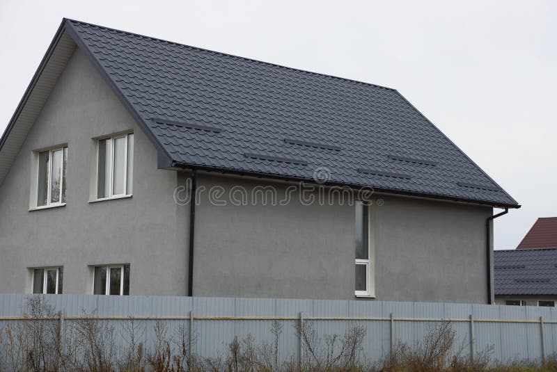Large gray private house with white windows under a brown tiled roof behind a metal fence stock image