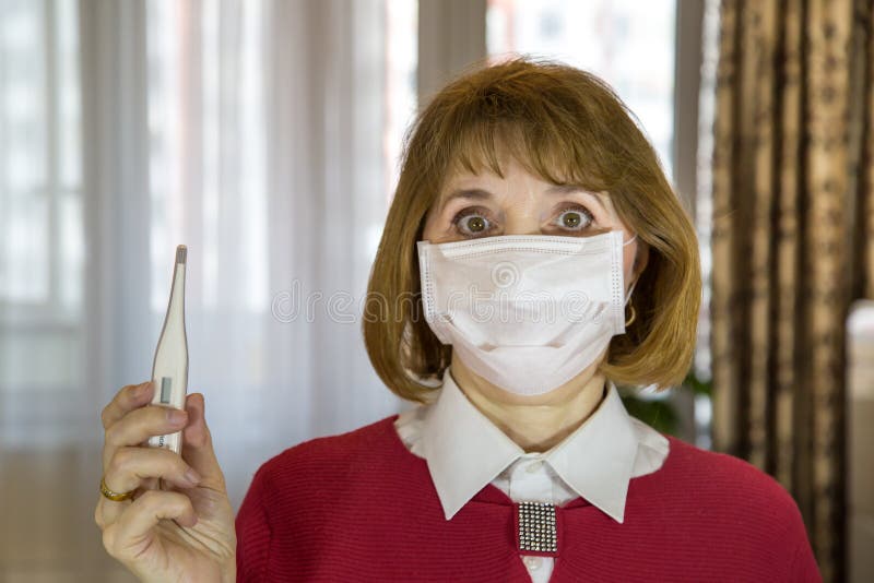 A middle-aged woman in a medical mask, looking at the camera and showing a thermometer in a room of an apartment building, protect royalty free stock photo