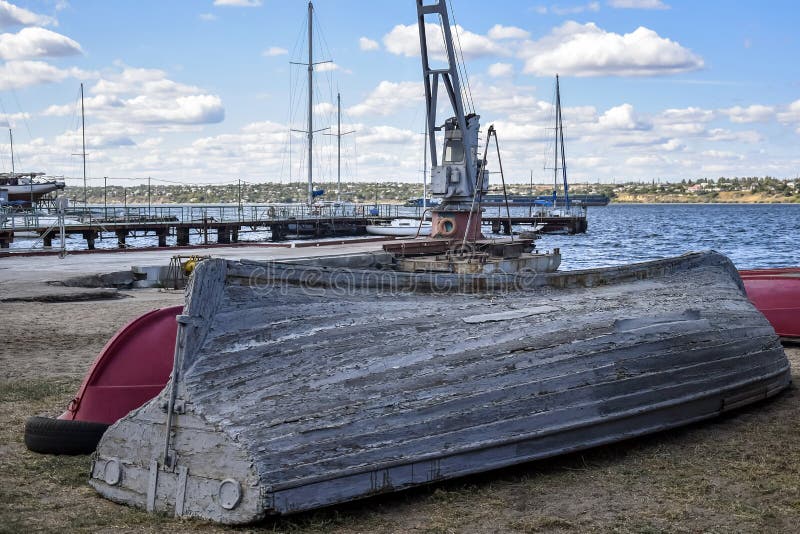 Old wooden boat is upside down on the river bank. Gray paint peels off the bottom and sides of the boat. View on the background of the river and the cloudy sky stock photo